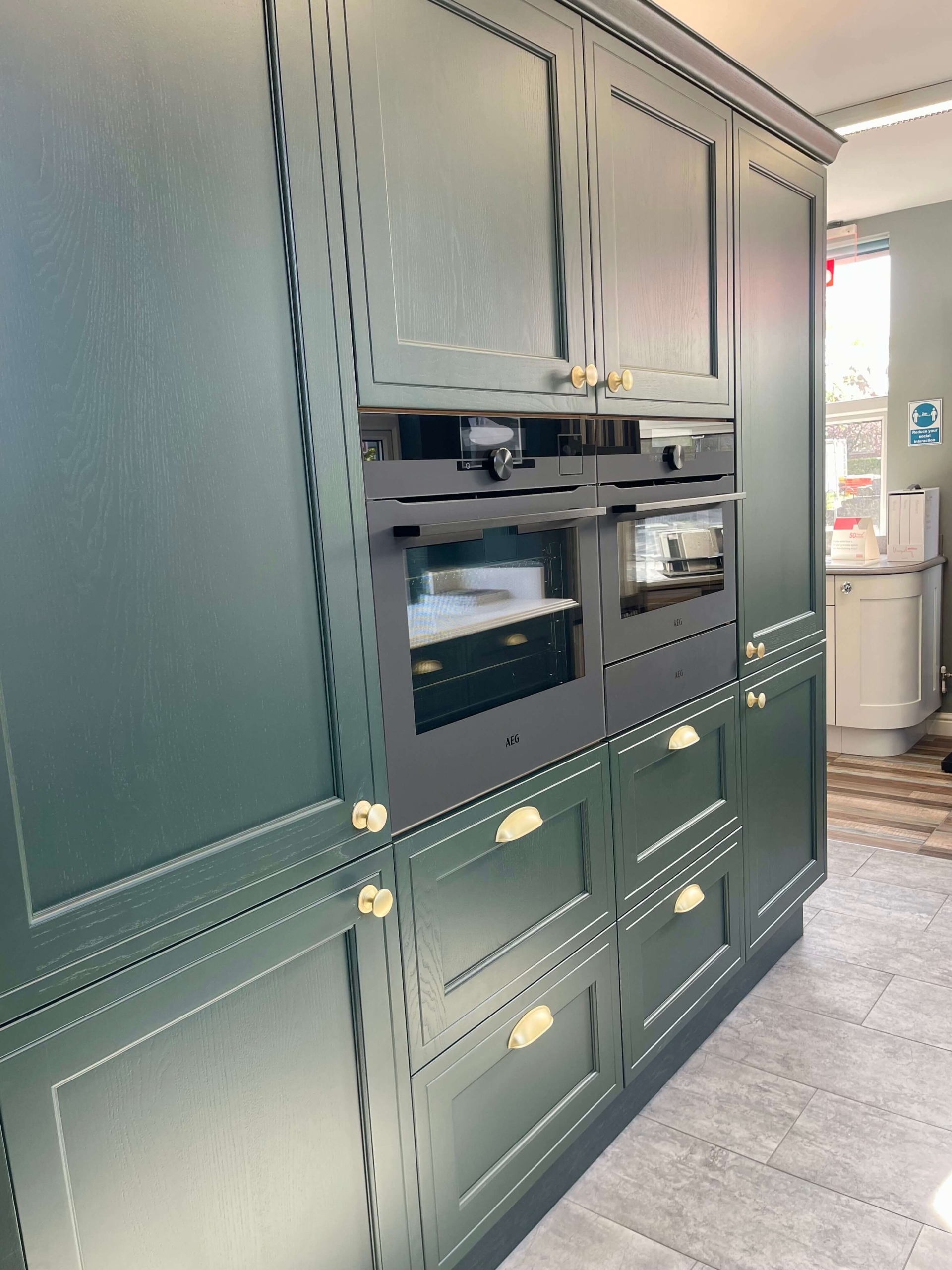 Teal high-gloss kitchen with island featuring marble-effect front, pendant lights, built-in ovens and wood-effect flooring at The Little Kitchen Factory showroom