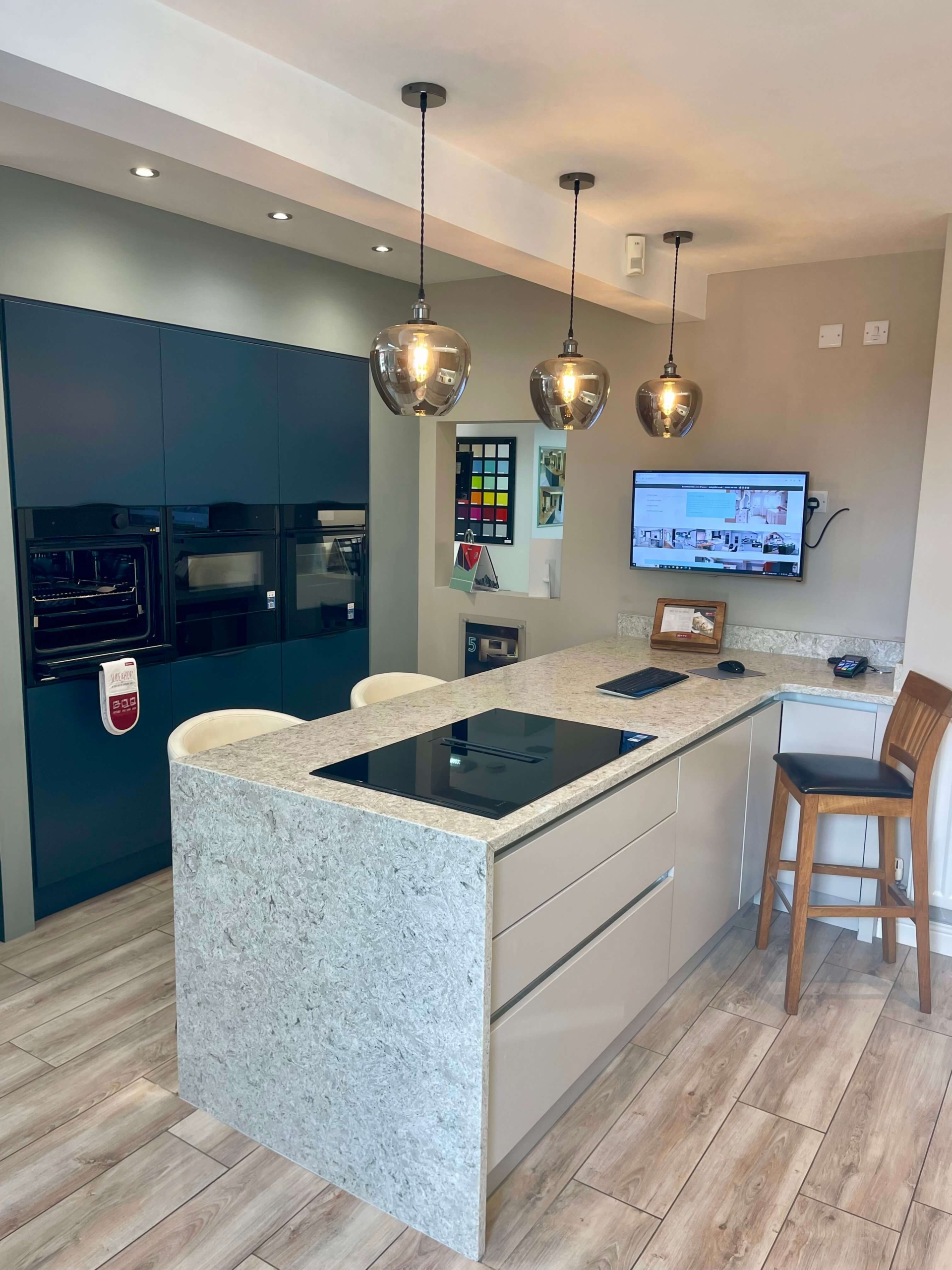 Teal high-gloss kitchen with island featuring marble-effect front, pendant lights, built-in ovens and wood-effect flooring at The Little Kitchen Factory showroom